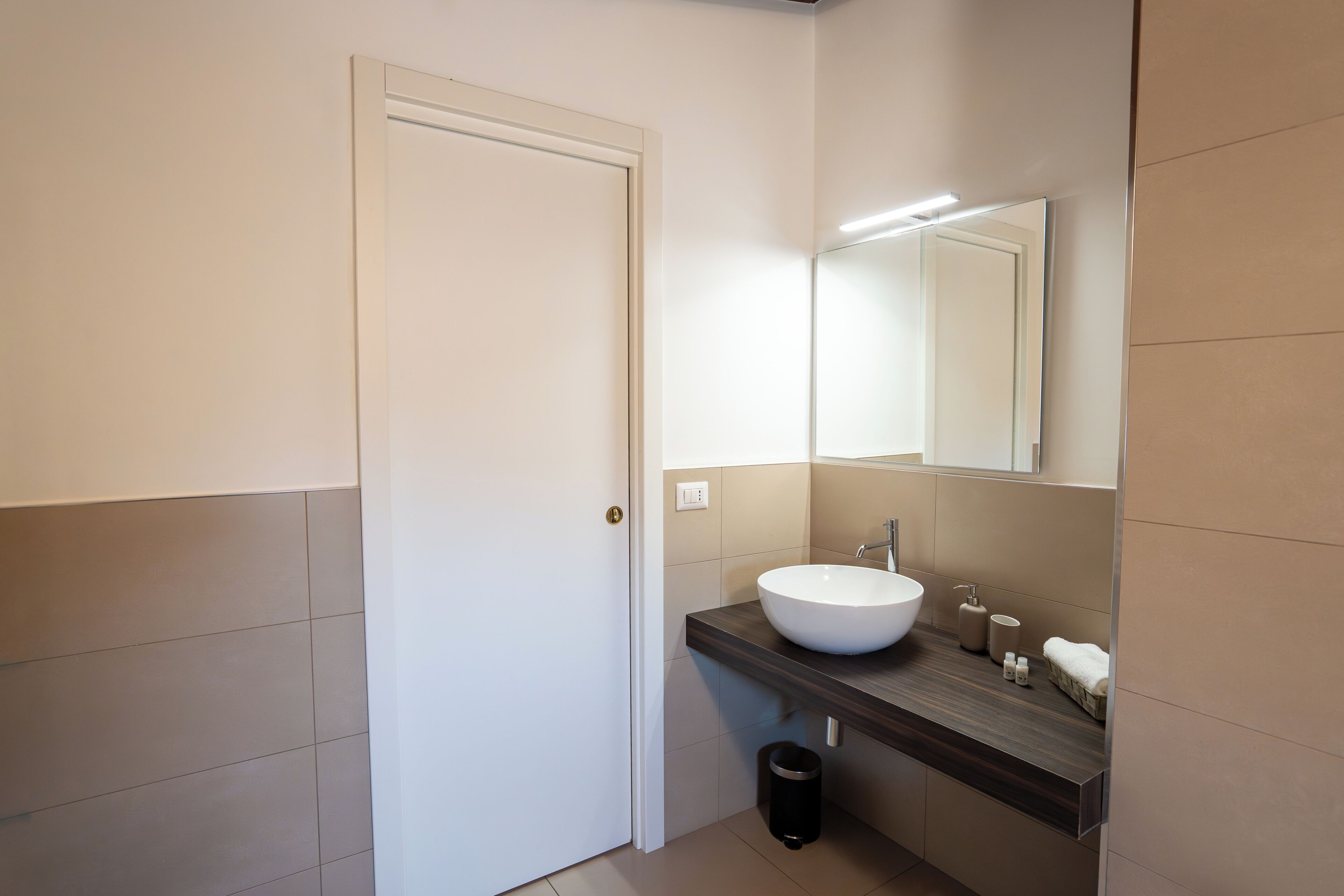 A bathroom with a white door, a large mirror, a white sink, a dark countertop, and a black trash bin.