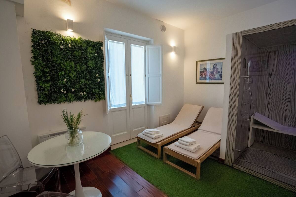 A room with a glass table, two lounge chairs, a green wall, and a shower area.