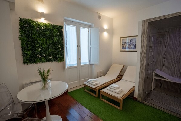 A room with a green wall, two lounge chairs, a small table with a vase, and a shower area.