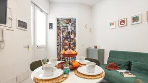 A small, well-lit room with a dining table set for two, a green sofa, and a colorful artwork on the wall.