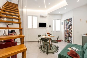 Ein modernes Wohnzimmer mit einer Holztreppe, einem Essbereich mit rundem Tisch und Stühlen, einem grünen Sofa und einem farbenfrohen Wandbild.
