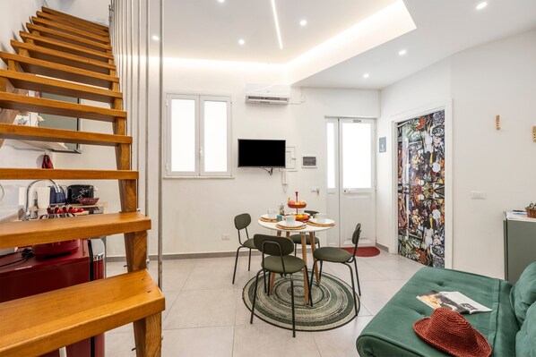Un espacio habitable moderno con una escalera de madera, una zona de comedor con una mesa y sillas redondas, un sofá verde y una pieza de arte colorido en la pared.