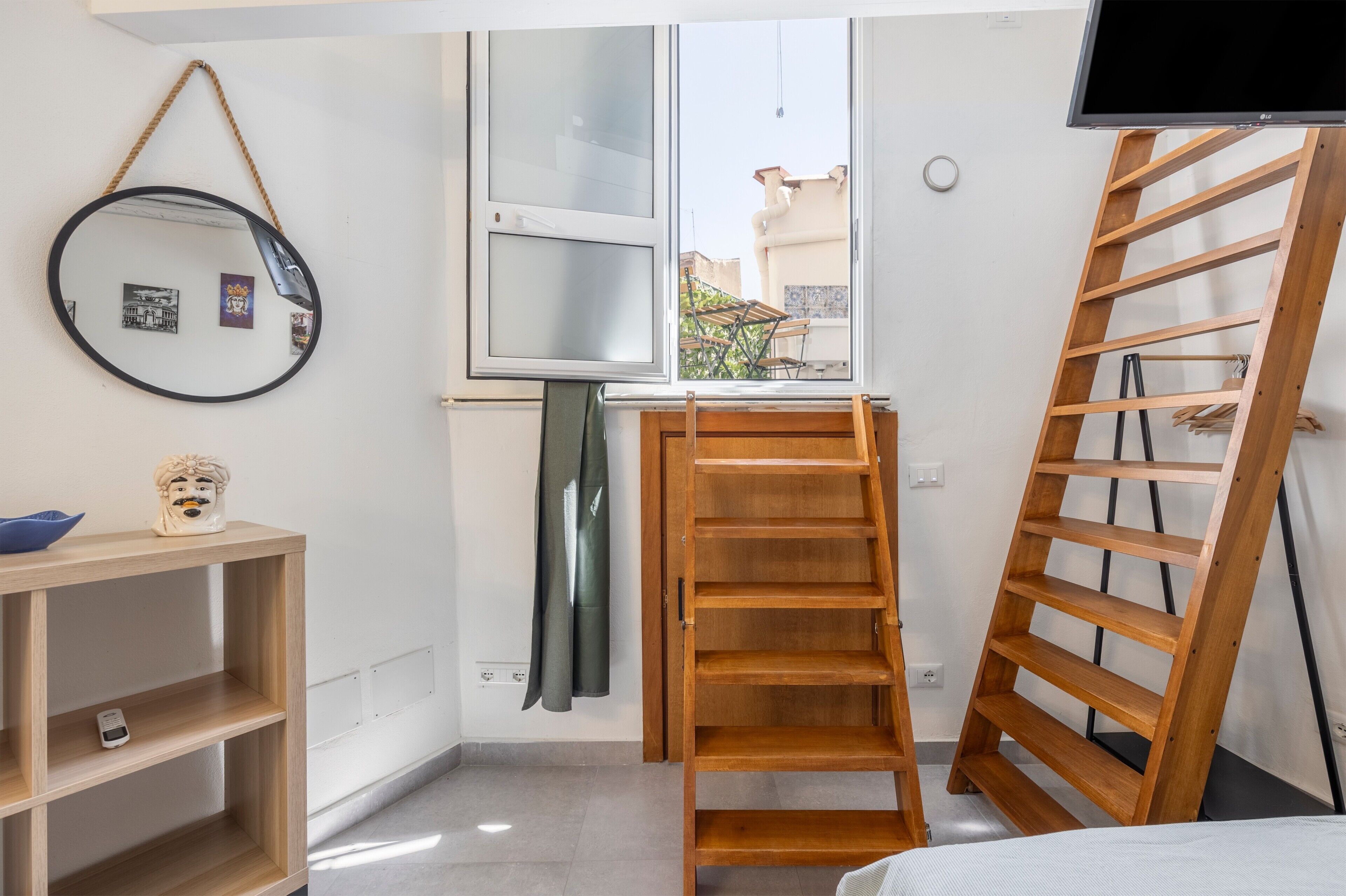 Ein Zimmer mit einer Holzleiter, einem Spiegel, einem Regal mit einer kleinen Figur und einem Fenster mit Blick auf Gebäude.