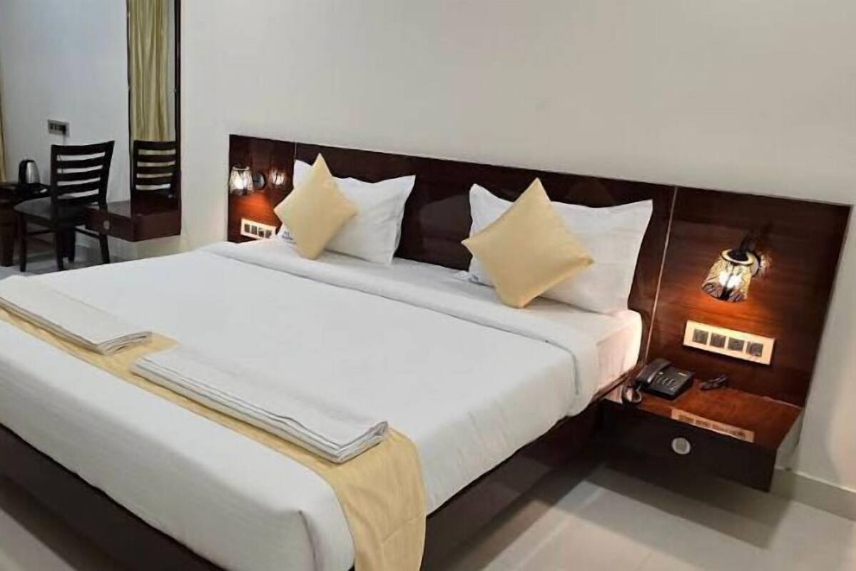 A neatly made bed with white bedding and two pillows, a bedside table with a lamp, and a chair in the background.