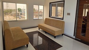 A spacious living room with a sofa and armchair, large windows offering a view of the outside, and a ceiling fan. - Hotel Itagi Square (Vijayapura)