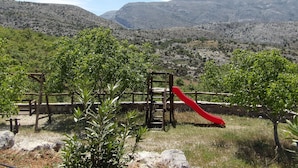 A playground with a red slide and climbing structure, surrounded by greenery and mountains in the background. - A traditional mountain gîte with swimming pool and local cuisine! (Κρουσώνας)