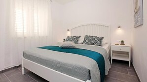 Ein Schlafzimmer mit einem Bett, weißen Nachttischen und einem Fenster mit durchsichtigen Vorhängen.