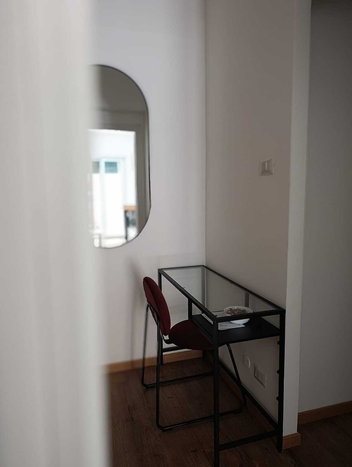 A small, modern room with a glass table and a red chair, a mirror on the wall, and a door leading to another room.