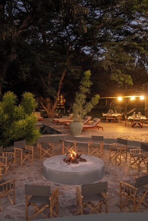 An outdoor seating area with a circular fire pit surrounded by wooden chairs, a large tree, and a dining area with tables and chairs. - Klaserie Drift Amani (Klaserie Private Nature Reserve)