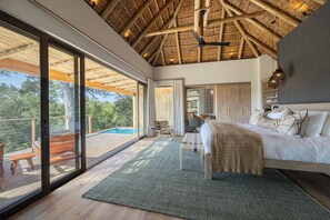 A bedroom with a large bed, a desk, a chair, and a view of a swimming pool. - Klaserie Drift Amani (Klaserie Private Nature Reserve)