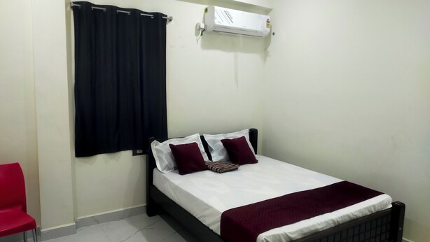 A single bed with a white and maroon bedspread, two pillows, a dark headboard, and a red chair in a room with white walls and a tiled floor. - SRI NAVANEETHA KRISHNA MAHAL & RESIDENCY (Andipatti)
