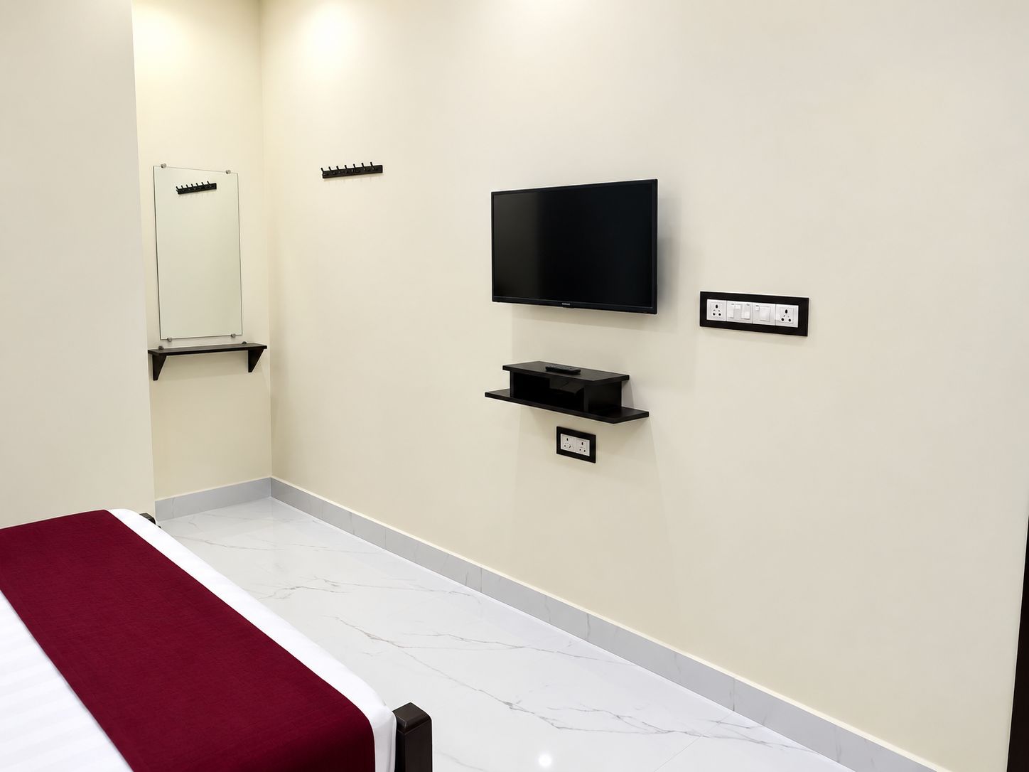 A hotel room with a bed, a television mounted on the wall, a small shelf below the TV, and a power outlet.