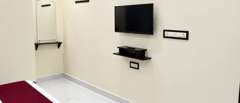 A hotel room with a bed, a television mounted on the wall, a small shelf below the TV, and a power outlet.