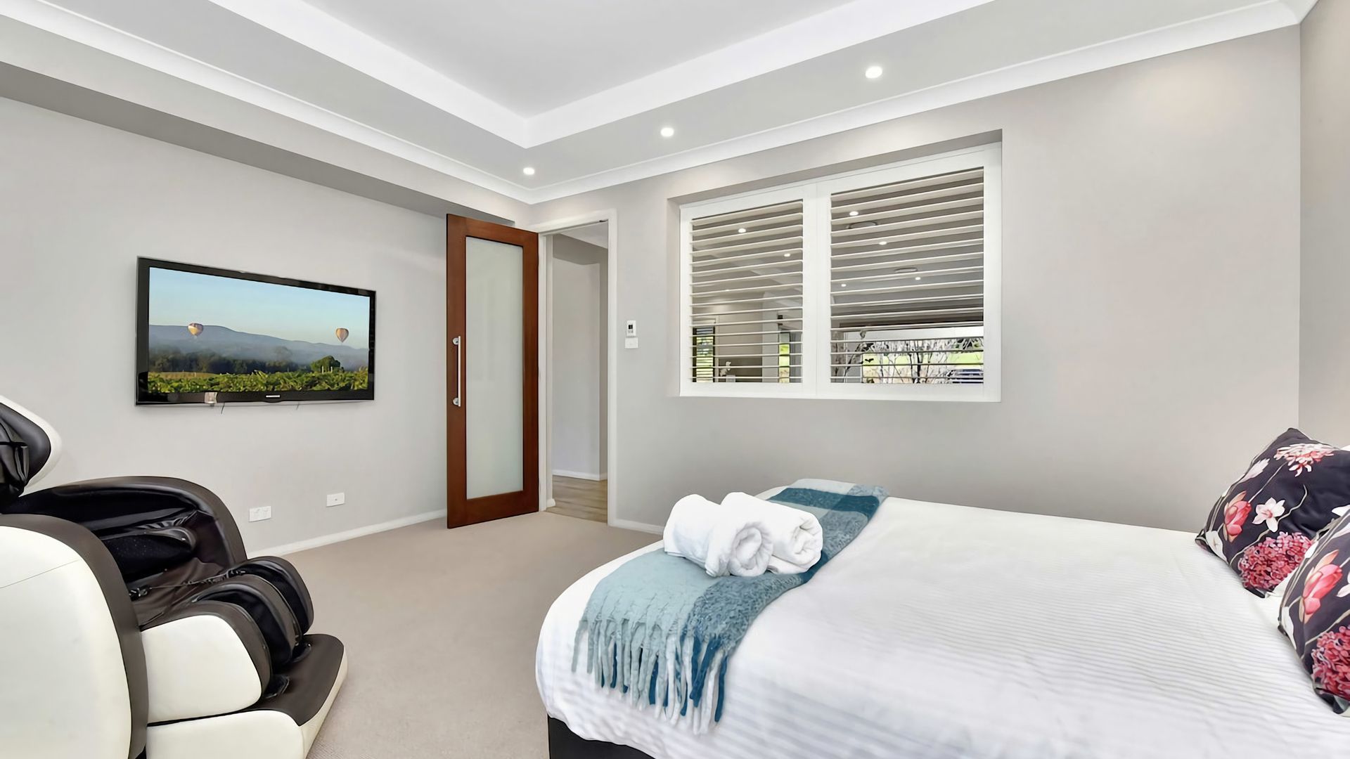 A modern bedroom with a bed, a television, a massage chair, and a window with blinds.