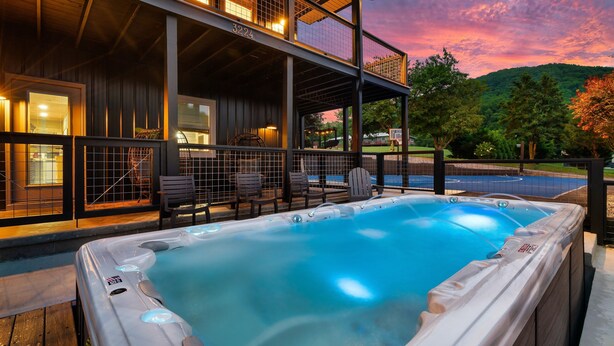 A hot tub with a wooden deck and chairs, a covered outdoor seating area, and a view of a mountain.