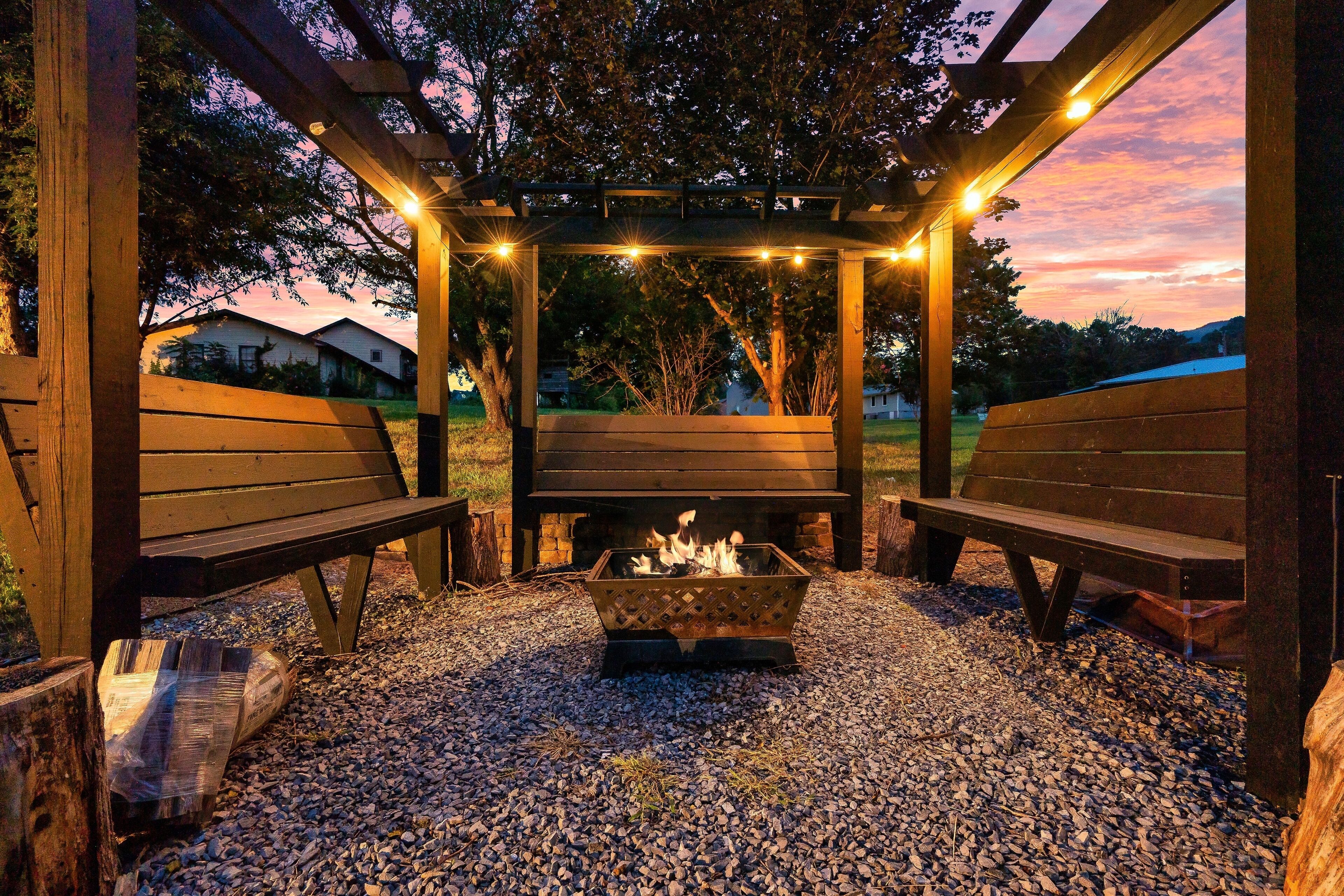 Une pergola en bois avec des bancs et un foyer, le tout illuminé par une guirlande lumineuse.