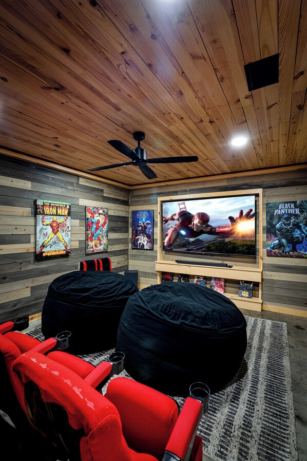 Una habitación con un televisor grande, dos sillones de mimbre, un ventilador de techo y carteles en la pared.