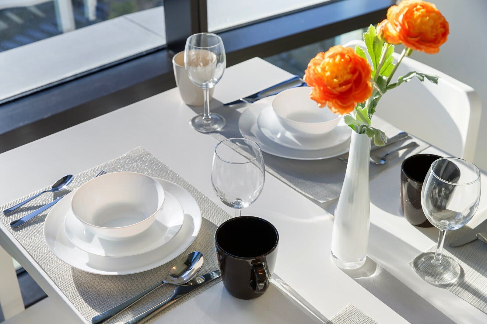 A table set for two with white plates, clear glasses, a black mug, and a vase with orange flowers.