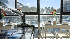 A piano in a dining area with a view of buildings through large windows.