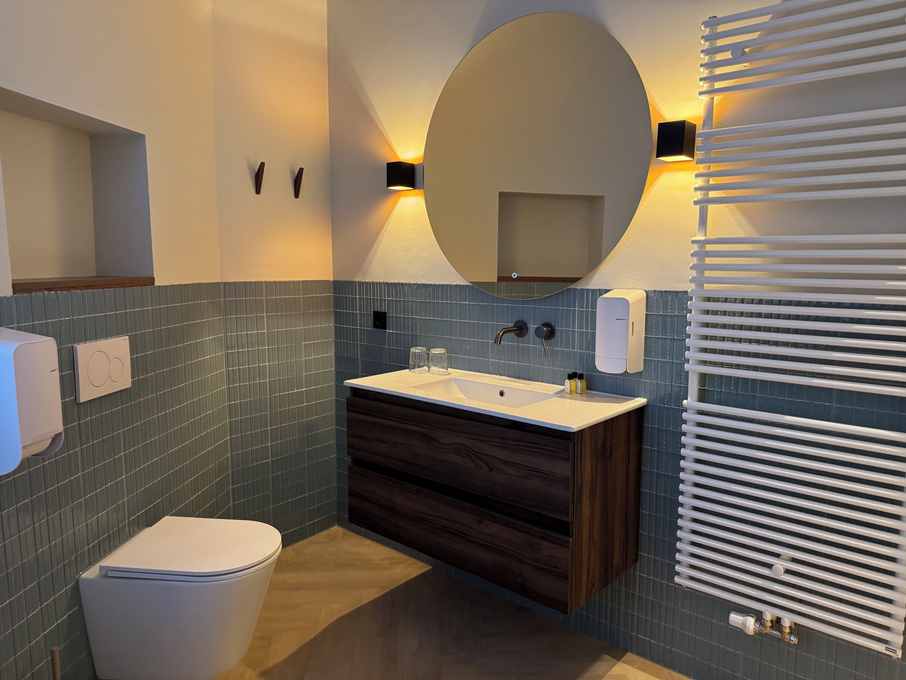 Une salle de bain moderne équipée de toilettes, d’un lavabo et d’un miroir. Les murs sont recouverts de carreaux bleus, le meuble-lavabo est en bois et le radiateur est blanc.
