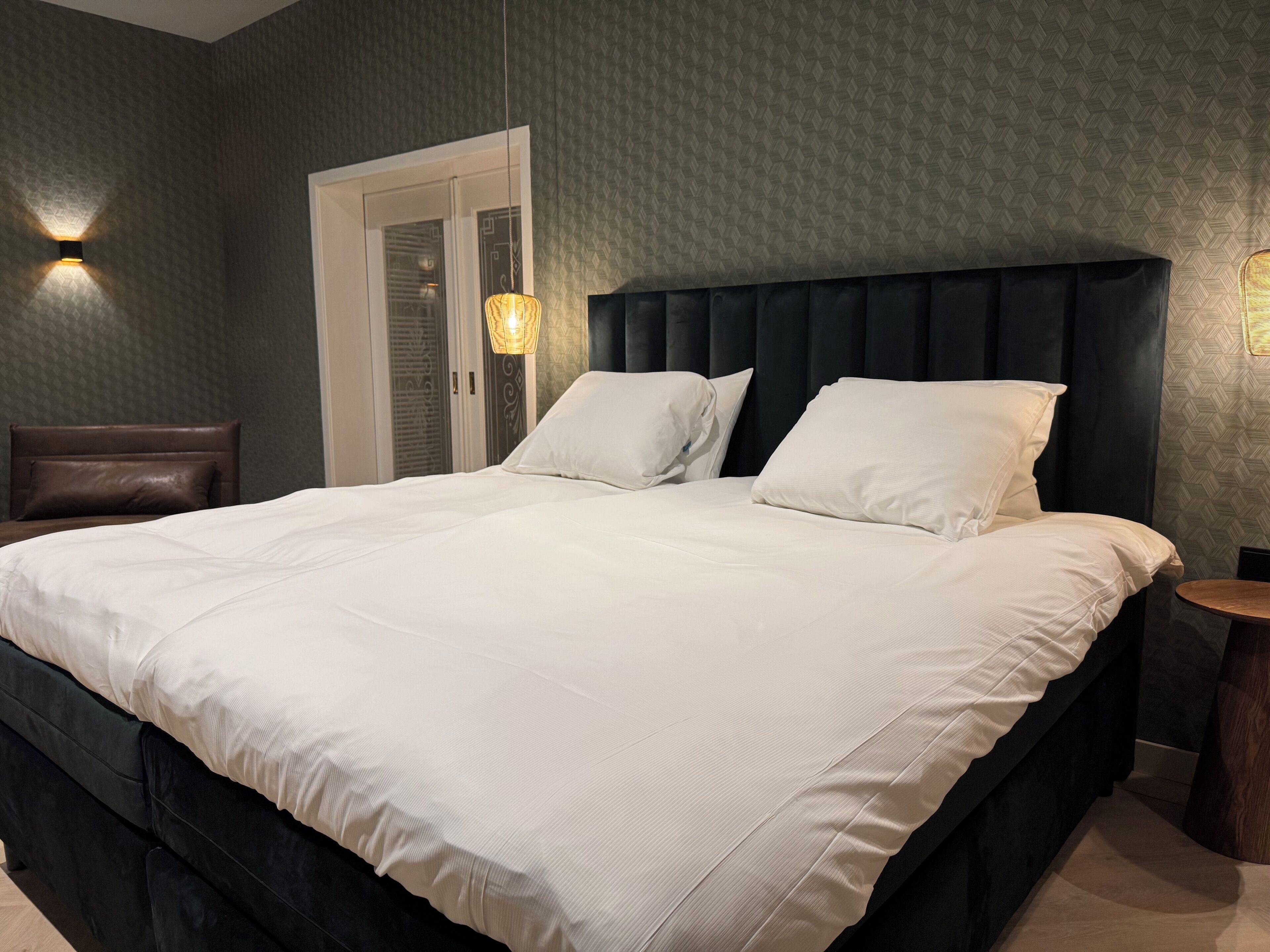 Un grand lit avec du linge de lit blanc, une tête de lit sombre et deux lampes fixées au mur.