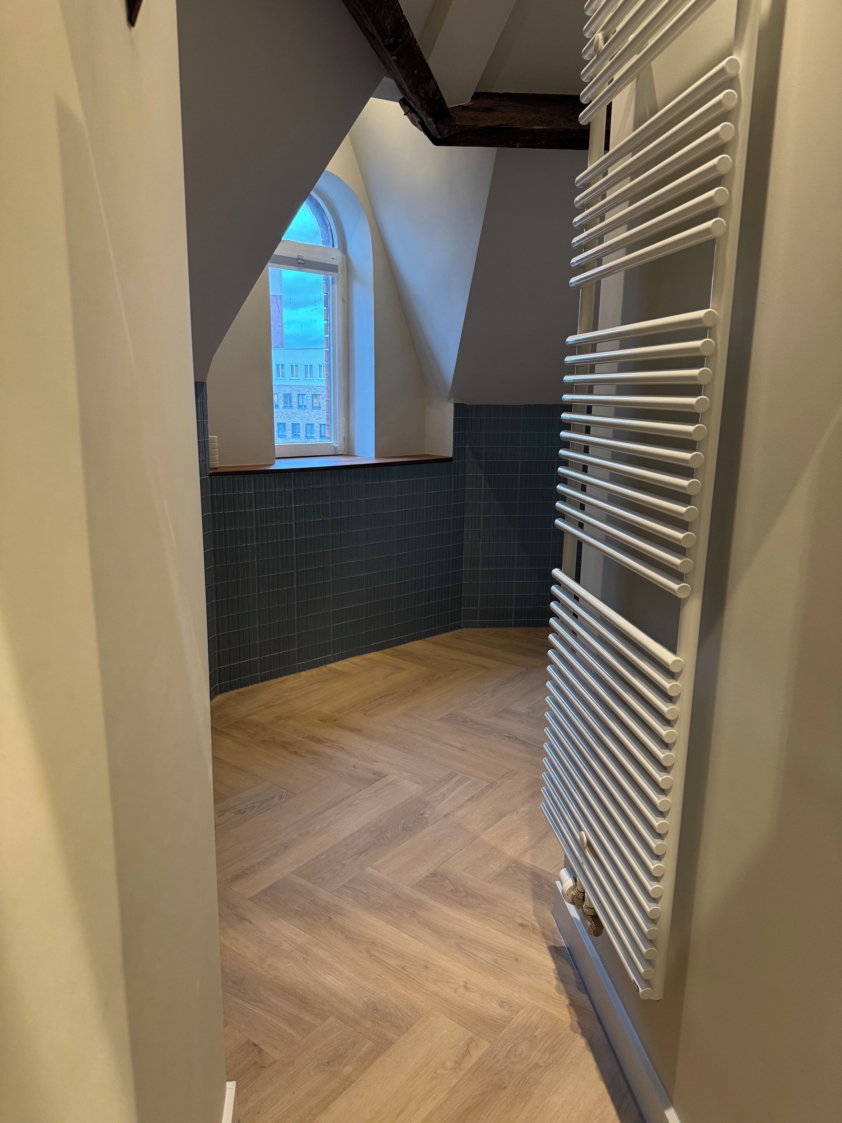 Een kamer met een schuine kap, een radiator, een raam met uitzicht op gebouwen en een visgraatvloer.