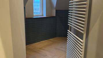 A room with a sloped ceiling, a radiator, a window with a view of buildings, and a herringbone-patterned floor.