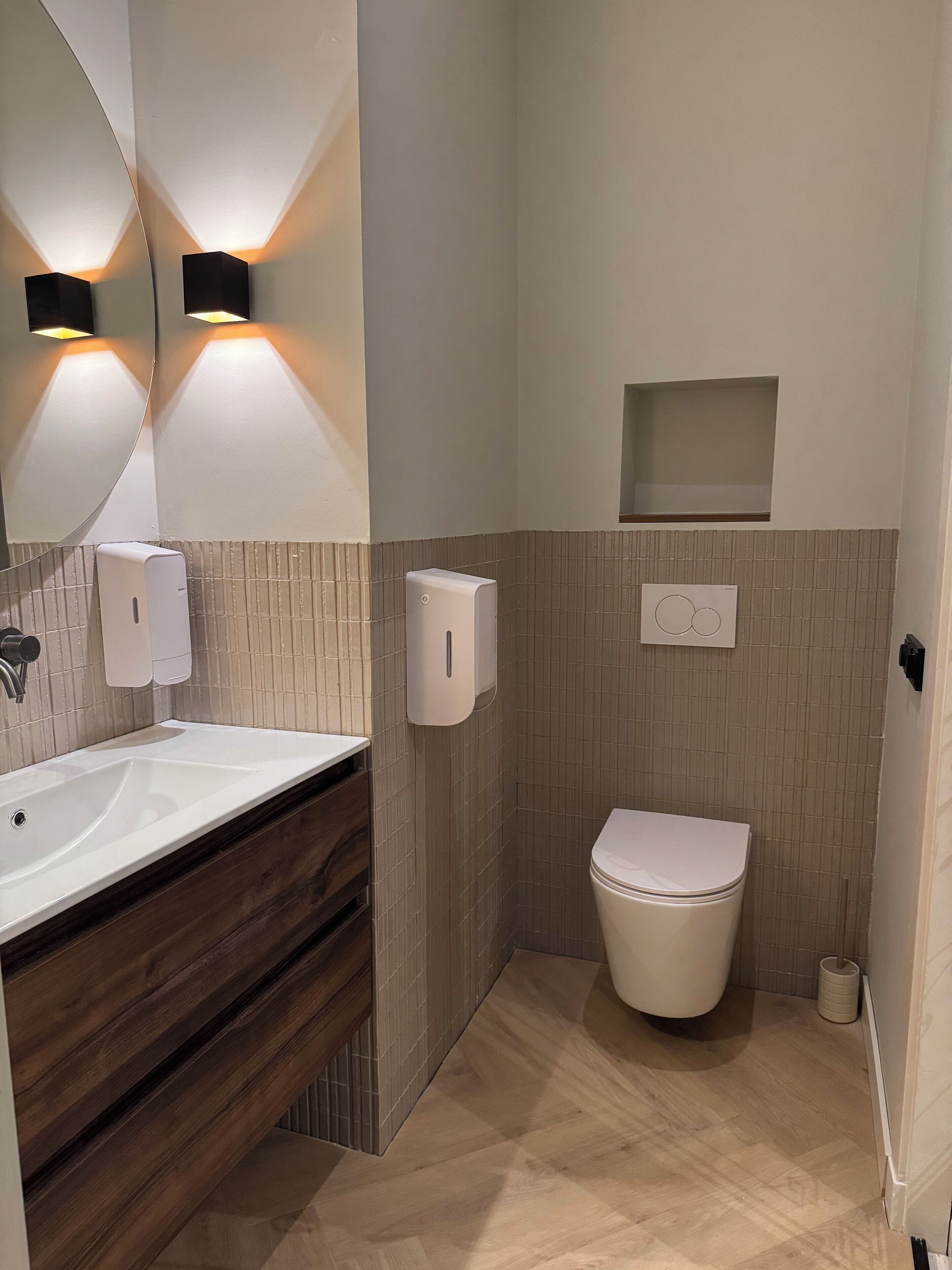 Un baño moderno con un lavabo blanco, tocador de madera, inodoro e iluminación empotrada en la pared sobre un espejo.