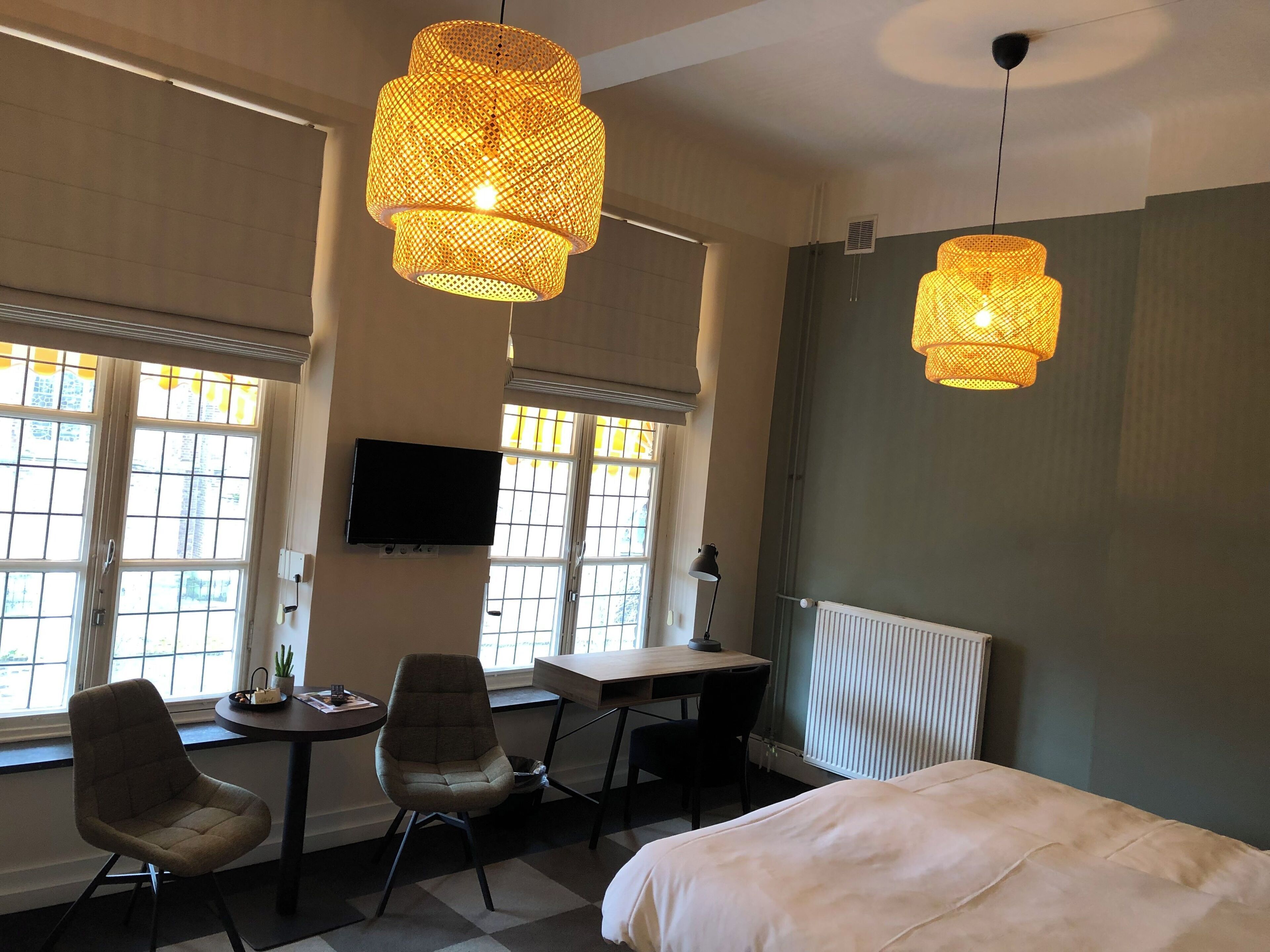 A hotel room with a bed, two chairs, a desk, a television, and two hanging lamps.
