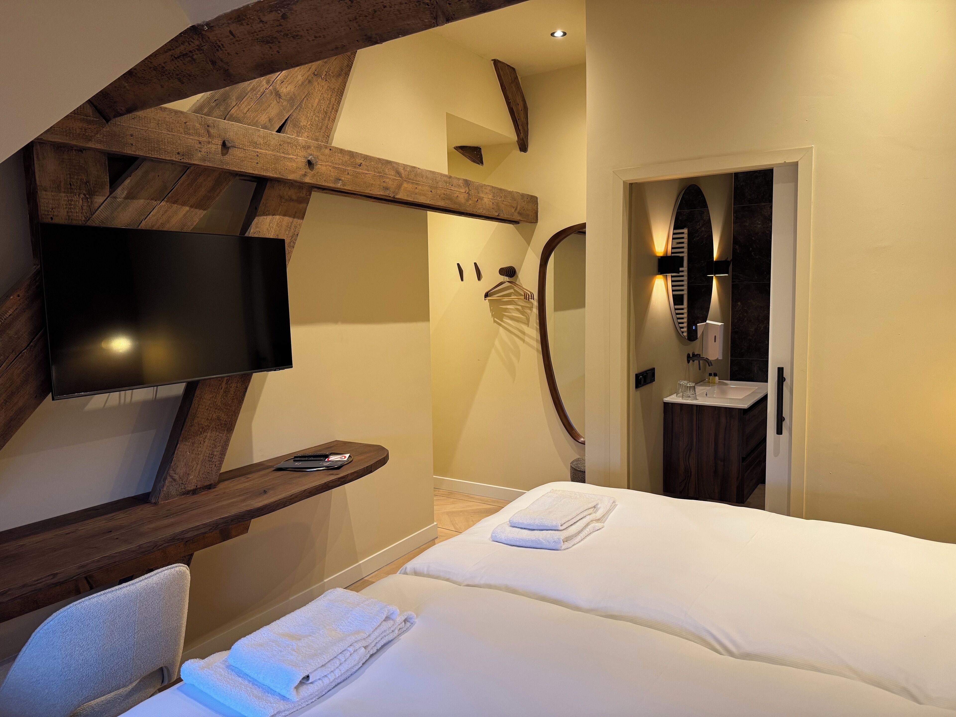 Una habitación de hotel moderna con una televisión de pantalla plana, un banco de madera, una cama con toallas, ducha y baño con espejo y lavamanos.