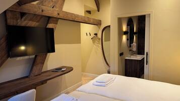 Una habitación de hotel moderna con una televisión de pantalla plana, un banco de madera, una cama con toallas, ducha y baño con espejo y lavamanos.