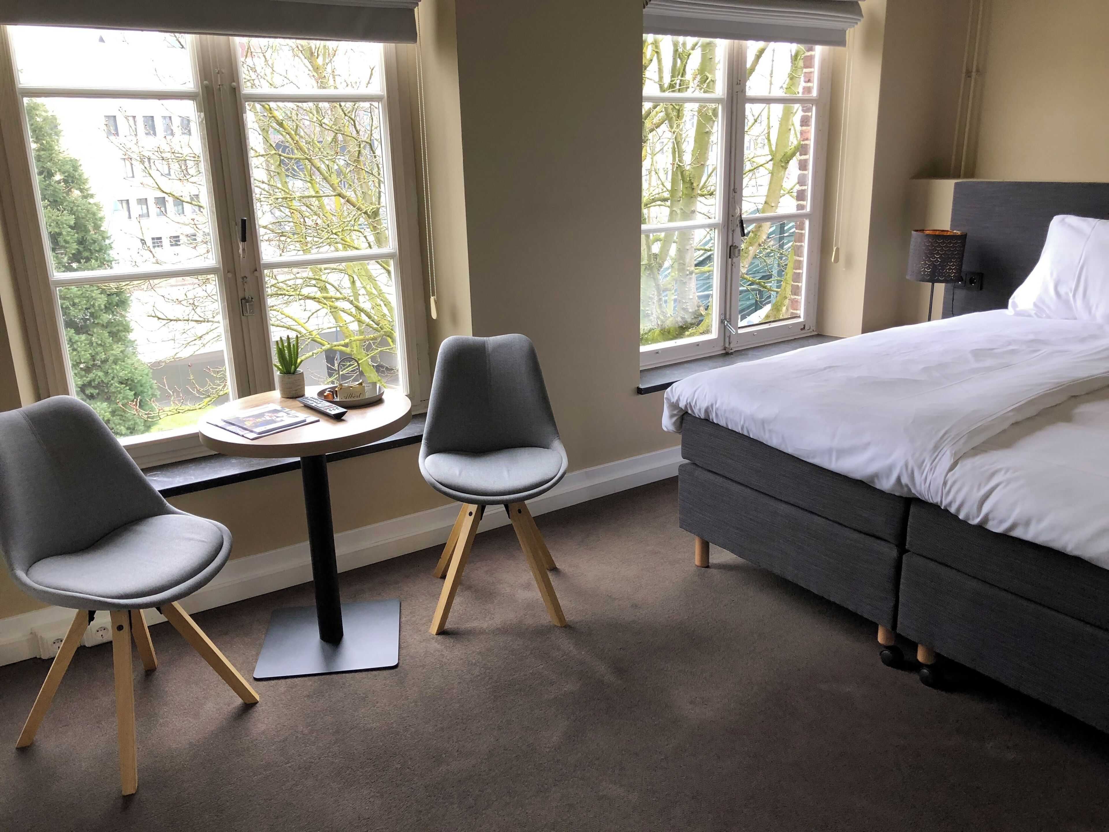 Ett hotellrum med en säng, två stolar, ett litet bord med en växt och stora fönster med utsikt över byggnader och träd.