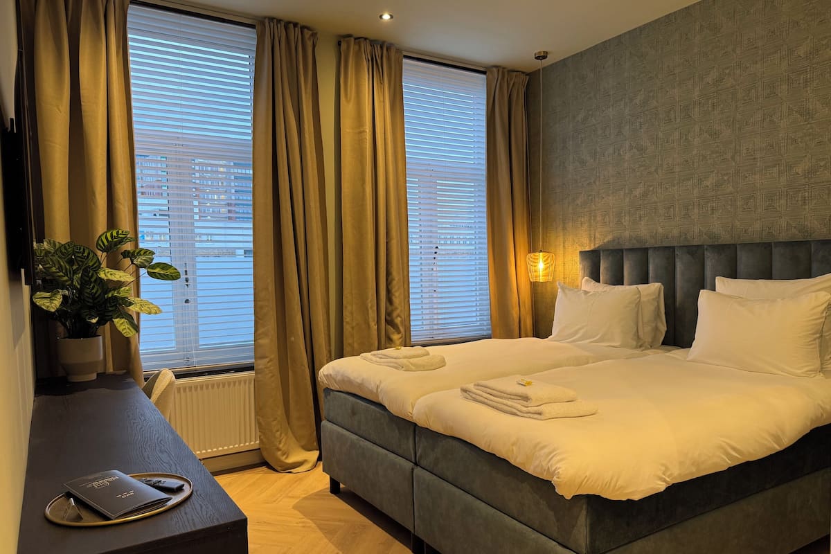 Habitación de hotel con una cama grande, una mesita de noche con una planta y vista a la ciudad desde la ventana.