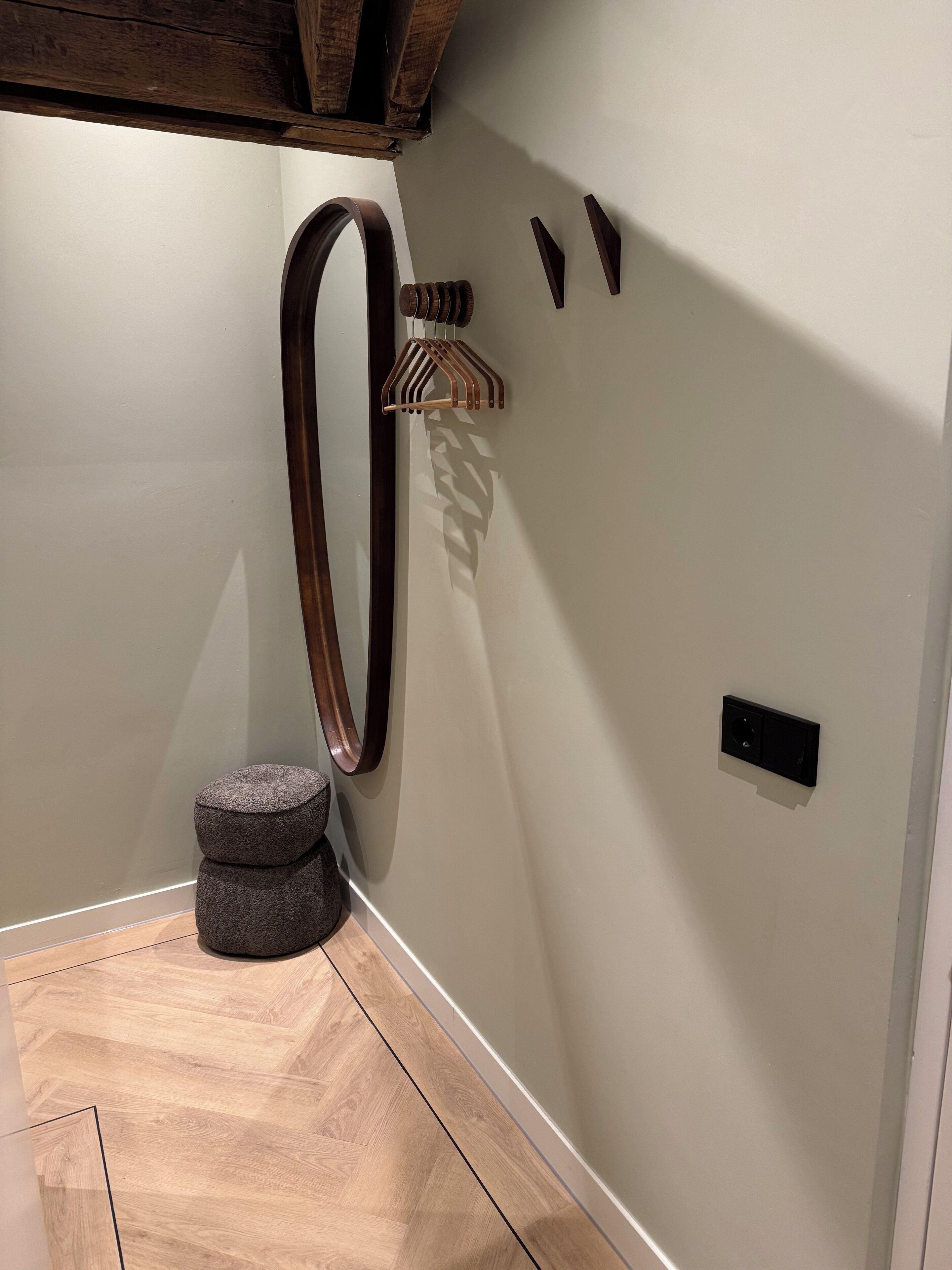 Een hoek van een kamer met een houten vloer, een grijze poef, een wandrek voor handdoeken en een klein zwart stopcontact.