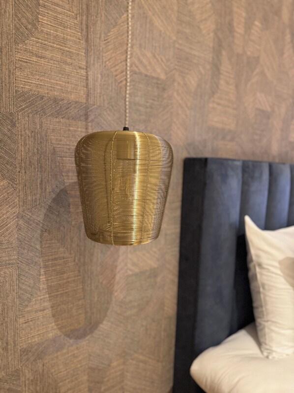 A hanging pendant light with a textured design, a patterned wall, and a bed with a dark headboard and white pillows. - De Pastorie Bed & Breakfast (Roermond)