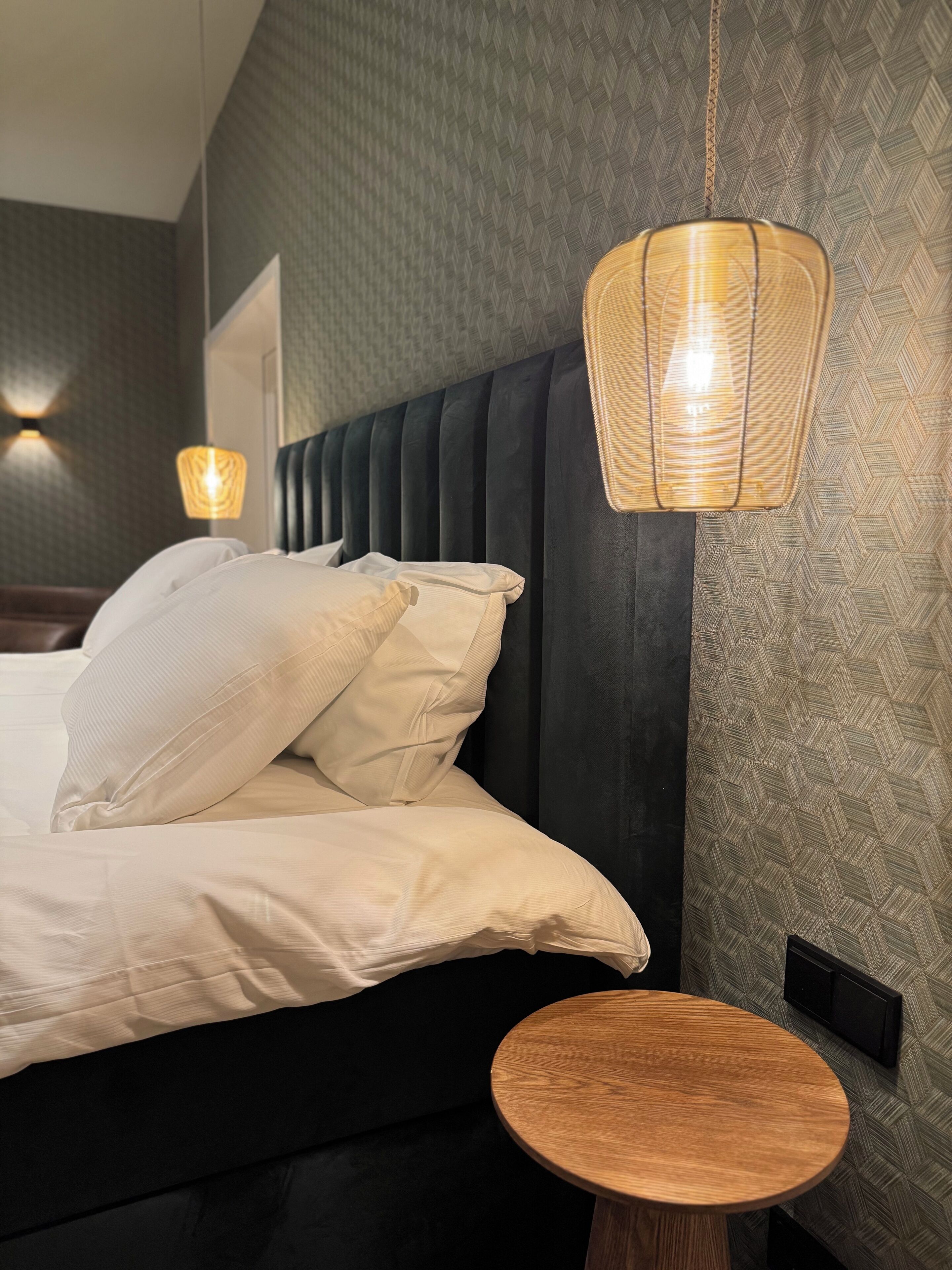 A neatly made bed with white linens and multiple pillows, a round wooden side table, and a hanging light fixture.