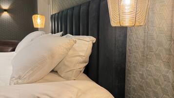 Un lit bien fait, avec du linge de lit blanc et plusieurs oreillers, une petite table d’appoint ronde en bois et un luminaire suspendu.
