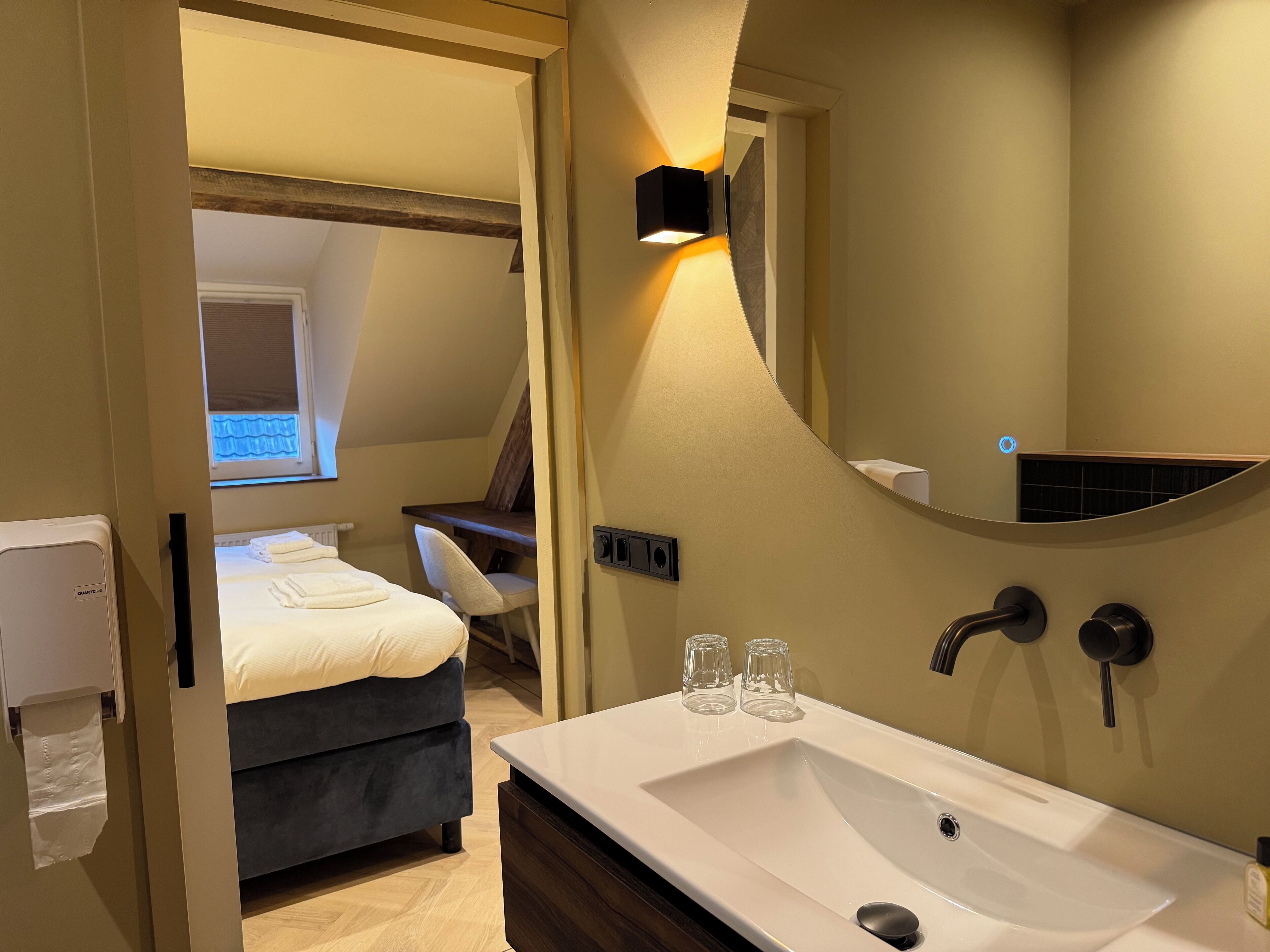 A modern bathroom with a large sink, a mirror, and a toilet. There is a bed with a window in the adjacent room.