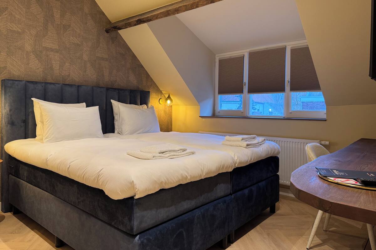 Un dormitorio con una cama grande, cabecera de madera, una ventana con persianas y una mesita con un libro.