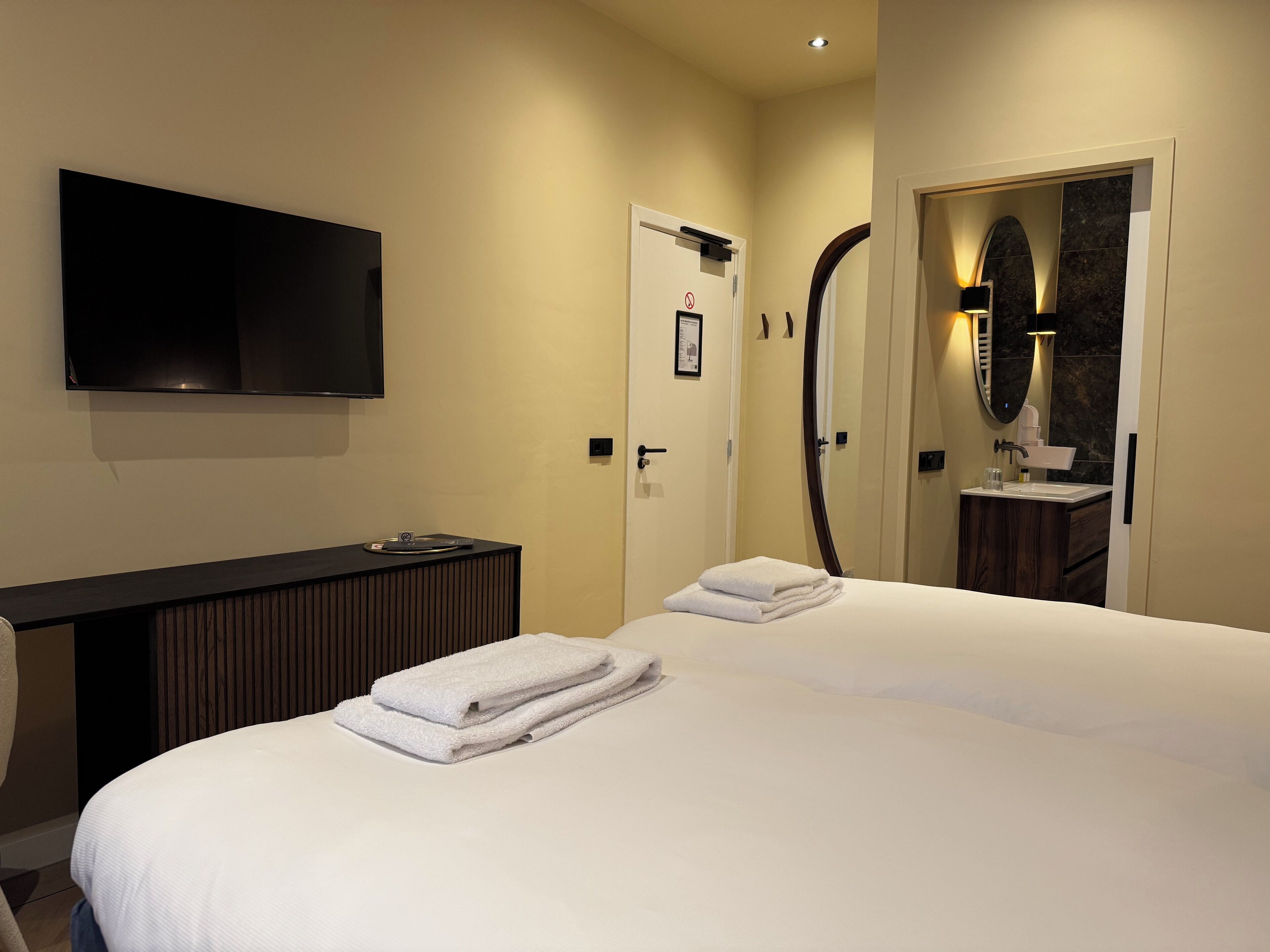 A hotel room with a bed, a TV mounted on the wall, a vanity with a mirror, and a door with a sign.