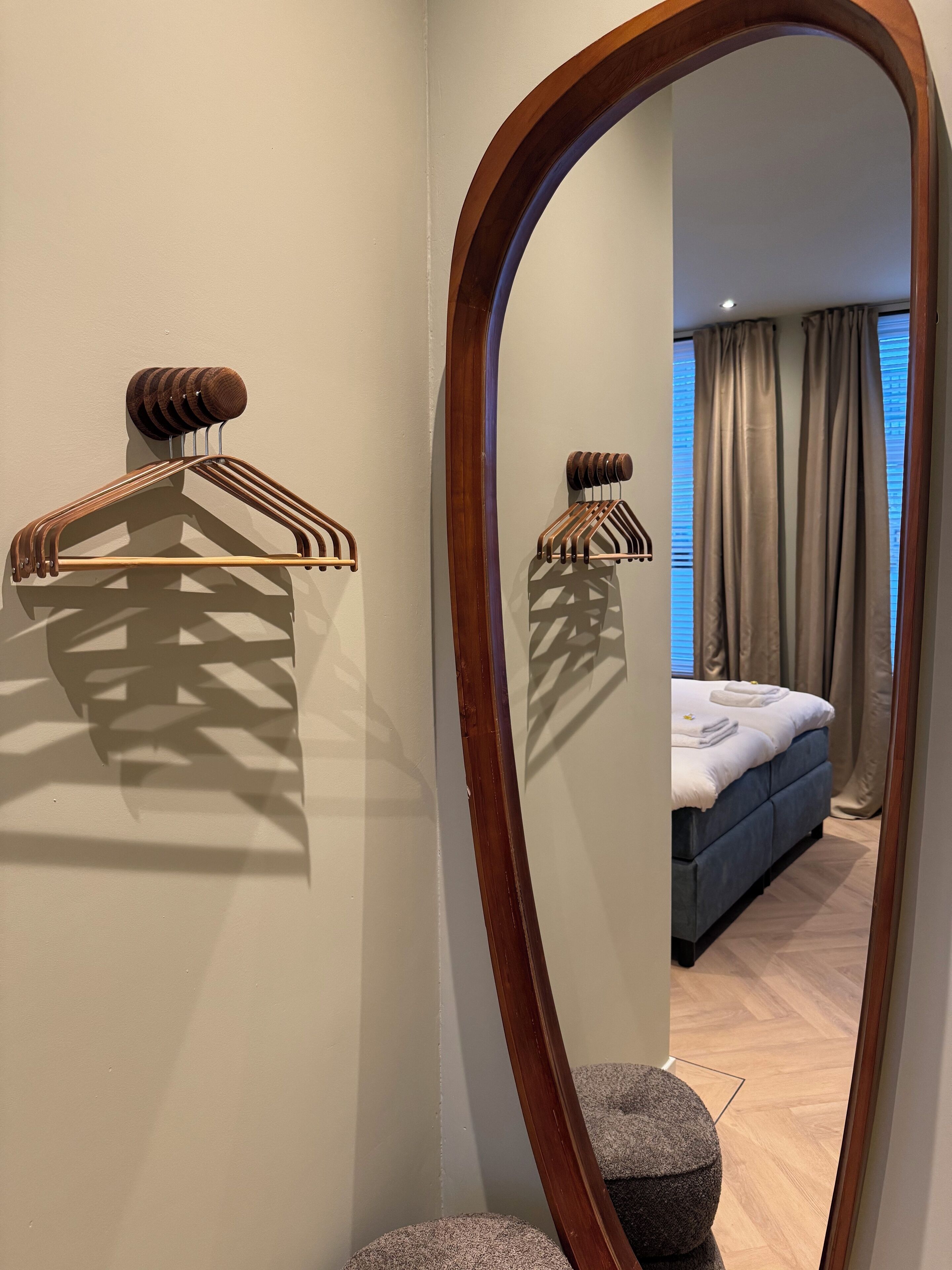 A mirror reflecting a room with a bed, curtains, and a wooden hanger on the wall.