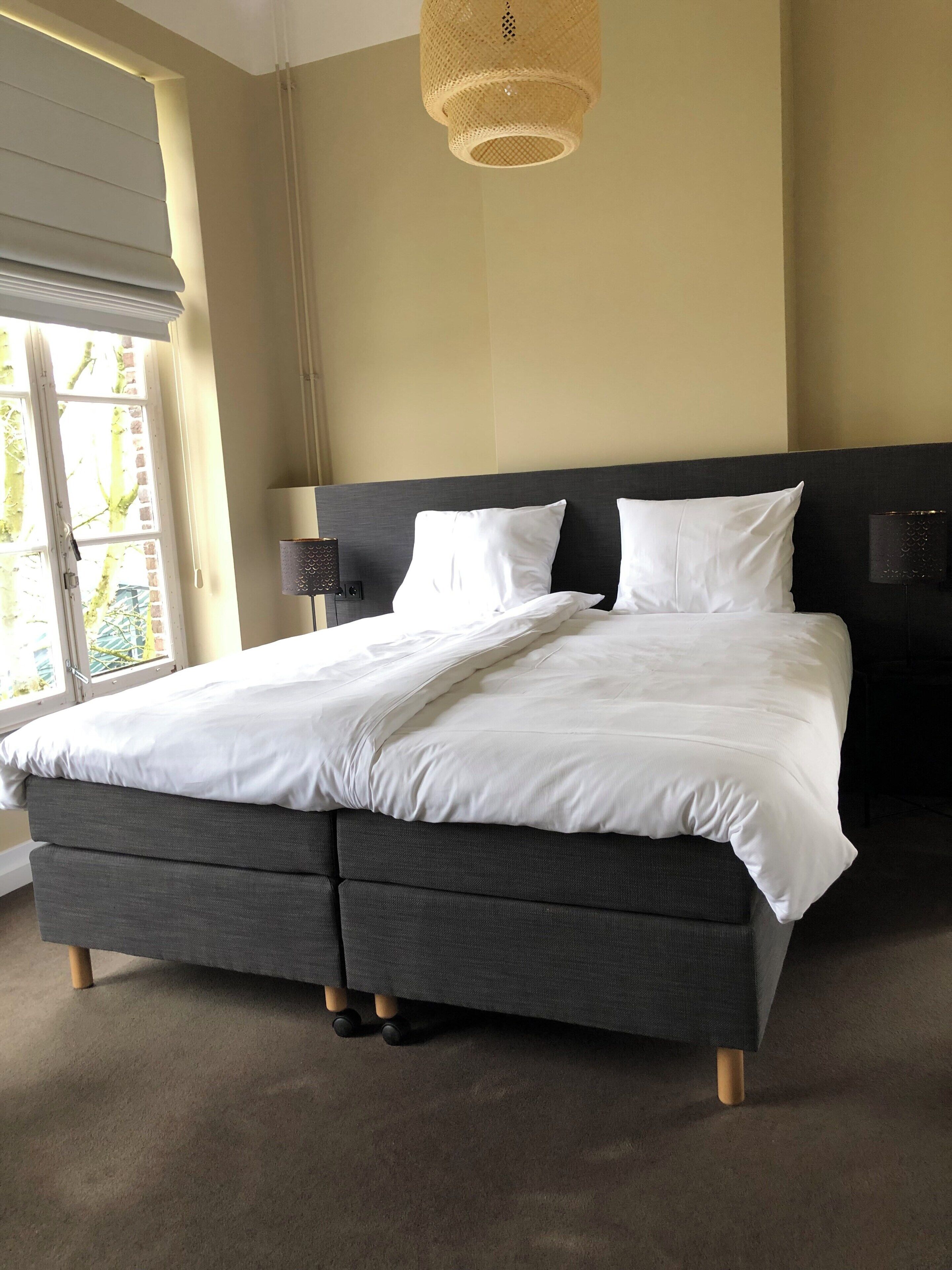 A modern bedroom with a large bed, two bedside tables, a hanging light fixture, and a window with blinds.