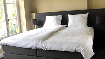 A modern bedroom with a large bed, two bedside tables, a hanging light fixture, and a window with blinds.