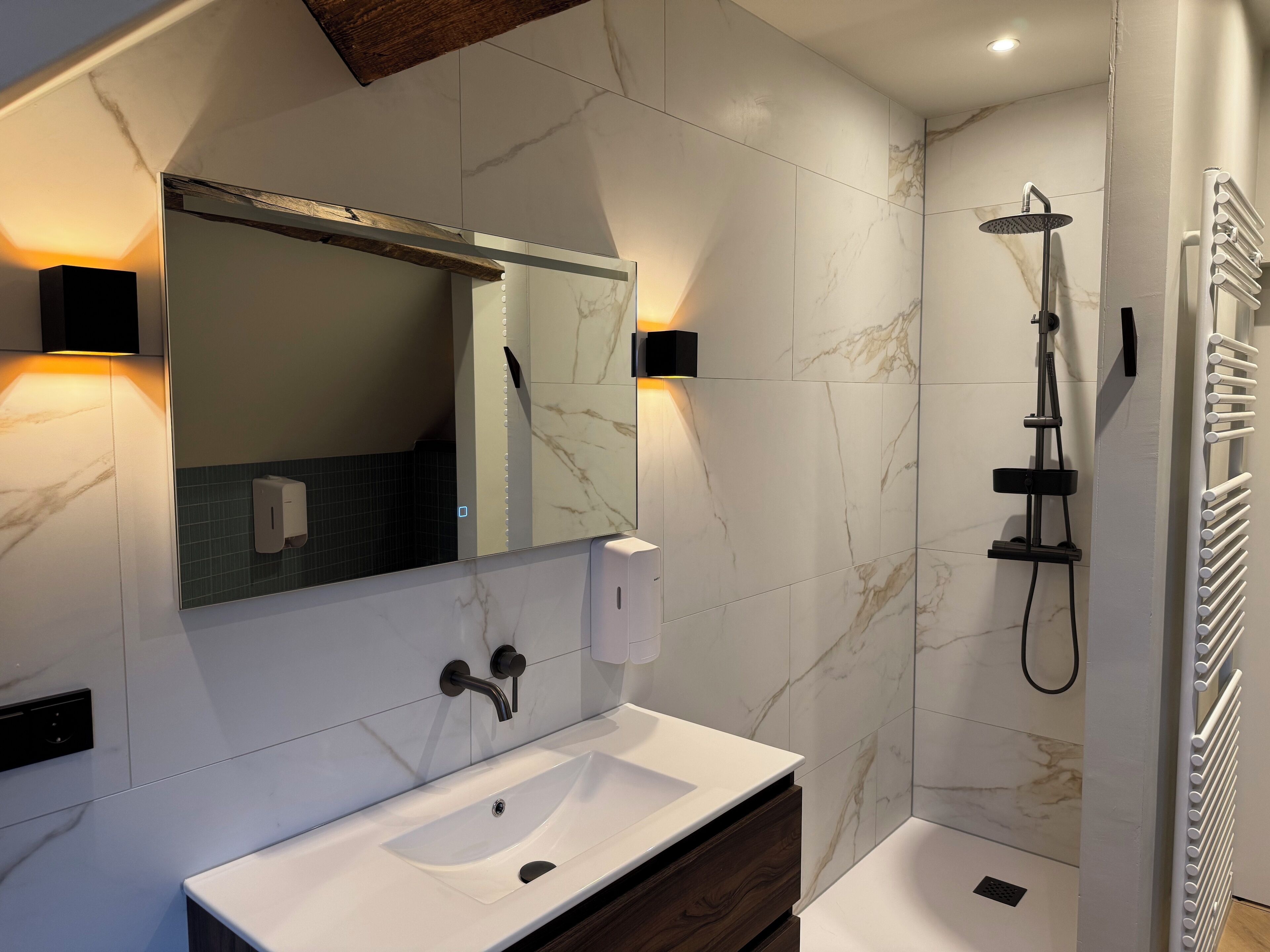 Une salle de bain moderne avec un lavabo blanc, un grand miroir, une douchette à main et un porte-serviettes.