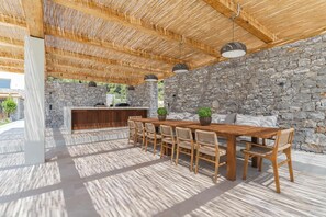 An outdoor dining area with a stone wall backdrop, wooden ceiling, and a stone fireplace. - Villa Delta Skiathos (Skiathos)