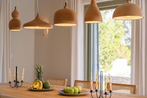 Un espace repas avec une table en bois, des fruits, des bougies et des lampes suspendues.