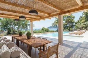 An outdoor dining area with a wooden table and chairs, a pergola with hanging lights, and a view of a pool and trees. - Villa Delta Skiathos (Skiathos)
