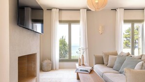 A modern living room with a fireplace, a sofa, a wooden table, and large windows offering a view of the sea. - Villa Delta Skiathos (Skiathos)