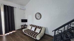Un salón con un sofá, un televisor sobre una mesita auxiliar, un aire acondicionado instalado en la pared y una escalera.