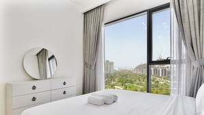 Une chambre moderne avec un grand lit, une commode à tiroirs, un grand miroir rond et une vue sur le paysage urbain, visible à travers de grandes fenêtres.